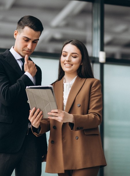 Dos personas de negocios revisando información en una tablet, con una mujer sonriendo y un hombre pensativo, en un entorno moderno de oficina.