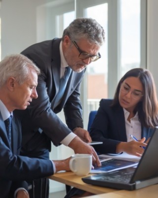 Reunión de negocios con tres profesionales discutiendo información en una laptop, con un hombre mayor señalando la pantalla y dos colegas atentos